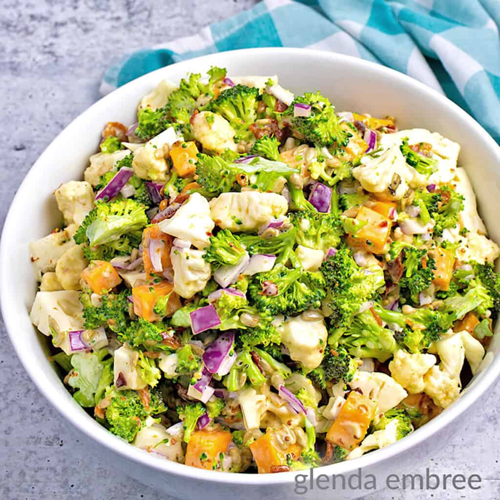 easy broccoli cauliflower salad recipe - salad in a round, white ceramic bowl ona concrete counter with a turquoise and white checked napkin
