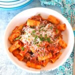 Rigatoni Bolognese served in a white stoneware bowl with low sides. A blue print fabric napkin and a fork are to the right of the bowl.